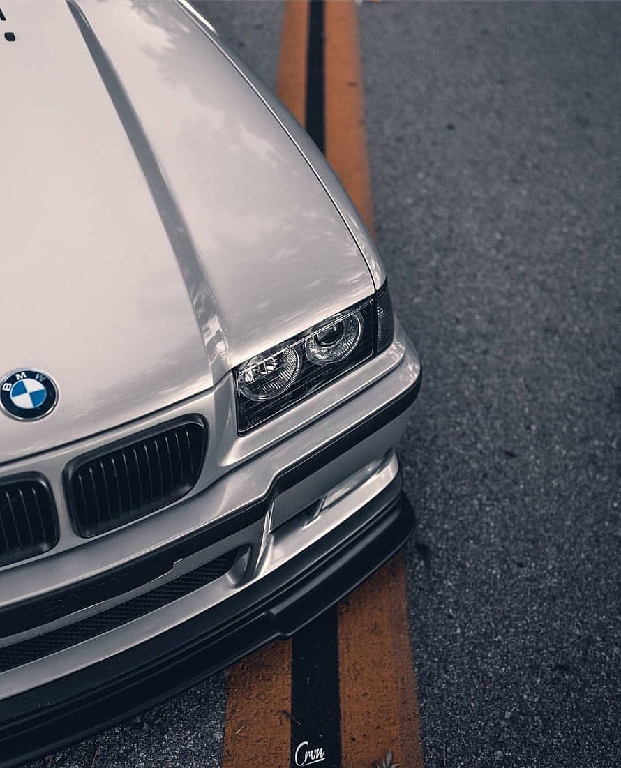 Close-up of a silver BMW E36 M3 front bumper and headlight on an asphalt road HD phone wallpaper