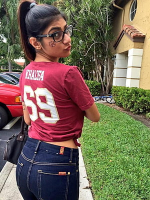 Mia Khalifa in a maroon jersey and dark denim jeans posing outdoors HD phone wallpaper
