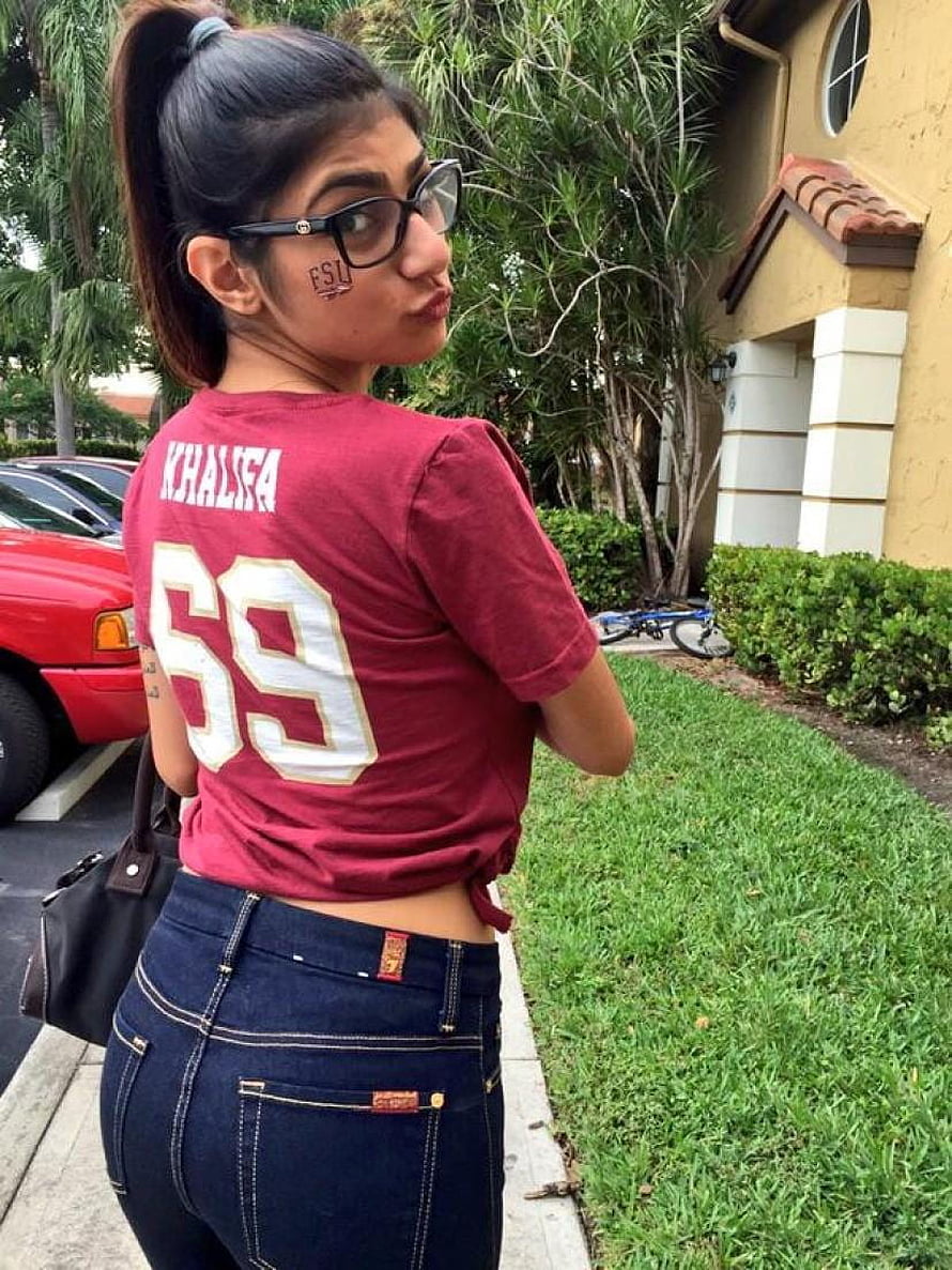 Mia Khalifa in a maroon jersey and dark denim jeans posing outdoors HD phone wallpaper