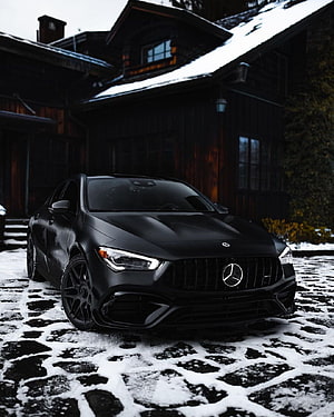 Matte Black Mercedes-AMG CLA 45 Parked on Snowy Cobblestones in Front of a Dark Wooden Chalet HD phone wallpaper