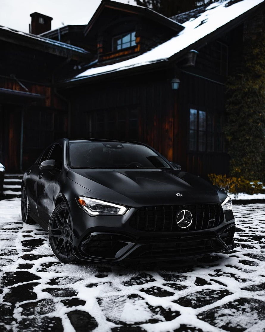 Matte Black Mercedes-AMG CLA 45 Parked on Snowy Cobblestones in Front of a Dark Wooden Chalet HD phone wallpaper