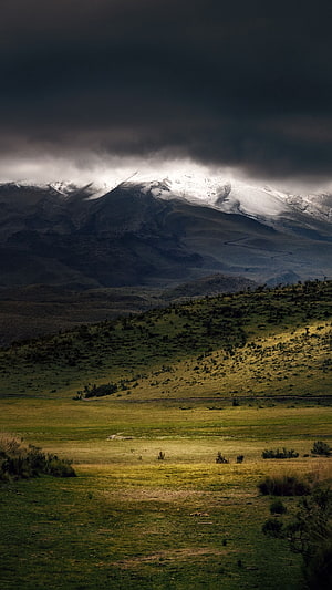 Dramatic Snow-Capped Mountain Peak Rising Above Green Rolling Hills Under Dark Storm Clouds HD phone wallpaper