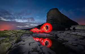 Vivid red light painting swirls on a rugged coastal cliff at twilight with a glowing reflection in a tide pool HD wallpaper