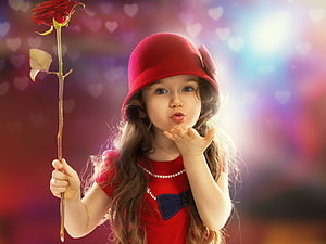 Adorable young girl in red hat blowing a kiss while holding a single red rose against a soft bokeh background HD wallpaper