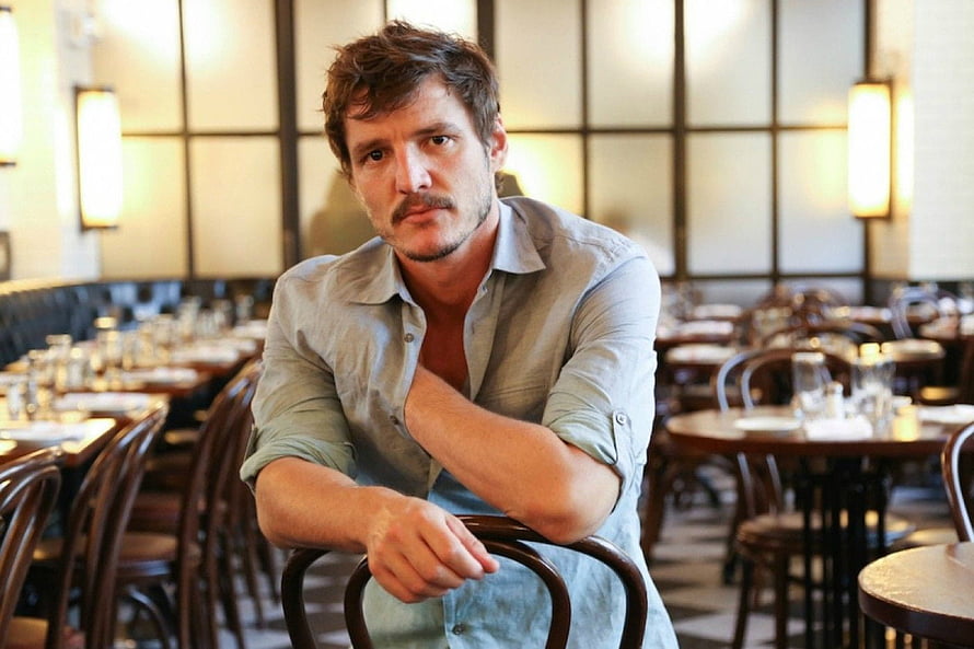 Pedro Pascal in a light shirt seated in a warmly lit restaurant with wooden chairs and tables HD wallpaper
