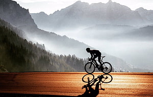 Silhouette of a cyclist riding a road bike across a golden field with misty mountains in the background HD wallpaper