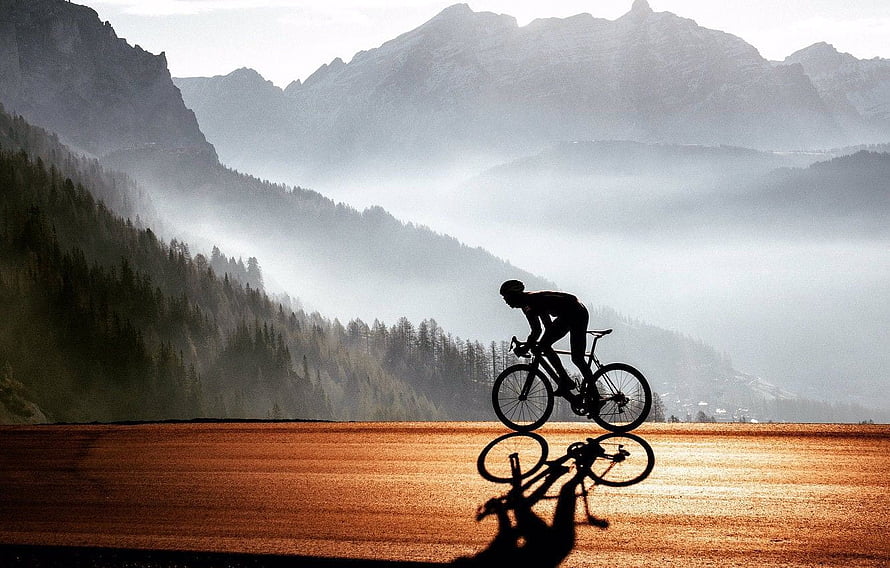 Silhouette of a cyclist riding a road bike across a golden field with misty mountains in the background HD wallpaper