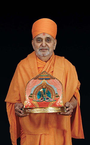 HH Pramukh Swami Maharaj in saffron robes holding a traditional deity portrait on a golden tray HD phone wallpaper