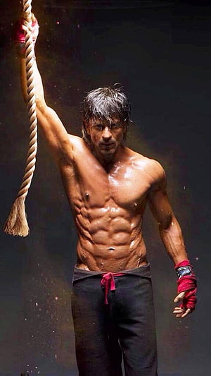 Shah Rukh Khan shirtless with defined abs gripping a rope in dramatic lighting HD phone wallpaper