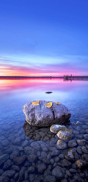 Serene Lake Sunset with Ginkgo Leaves on a Rock and Distant Pier HD phone wallpaper