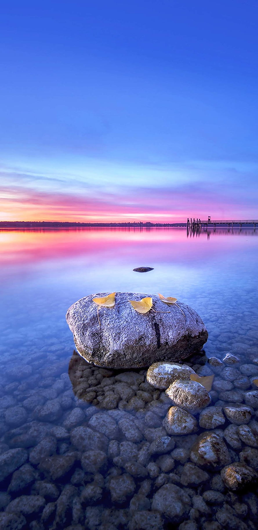 Serene Lake Sunset with Ginkgo Leaves on a Rock and Distant Pier HD phone wallpaper
