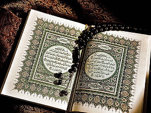 Open Quran with intricate green floral borders and black prayer beads resting on ornate fabric HD wallpaper
