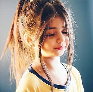 Portrait of a young girl with long brown hair in a ponytail, wearing a yellow shirt with blue trim, looking down with a gentle smile in soft lighting HD wallpaper