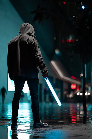 Mysterious hooded figure holding a glowing blue lightsaber on a rainy cyberpunk city street at night HD phone wallpaper