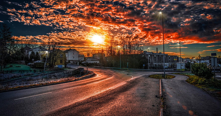 Dramatic Sunset Over a Winding Road with Fiery Orange Clouds and Suburban Buildings HD wallpaper