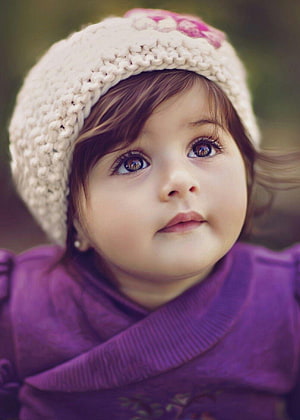 Adorable baby girl wearing a cream knit hat and purple top with a soft outdoor background HD phone wallpaper