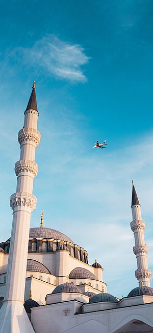 Airplane flying over mosque minarets and domes against blue sky HD phone wallpaper