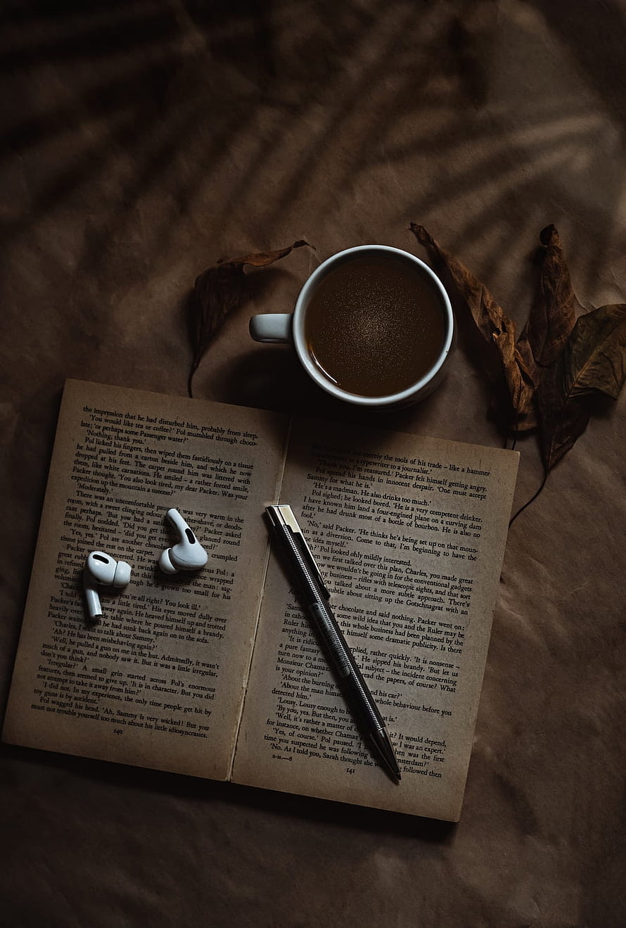 Cozy Reading Setup With Open Book, Coffee Cup, Wireless Earbuds, And Pen On Brown Fabric HD phone wallpaper