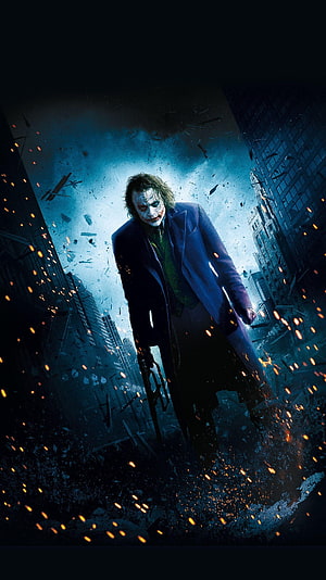 The Joker from The Dark Knight standing in a destroyed city street with flying debris and embers HD phone wallpaper