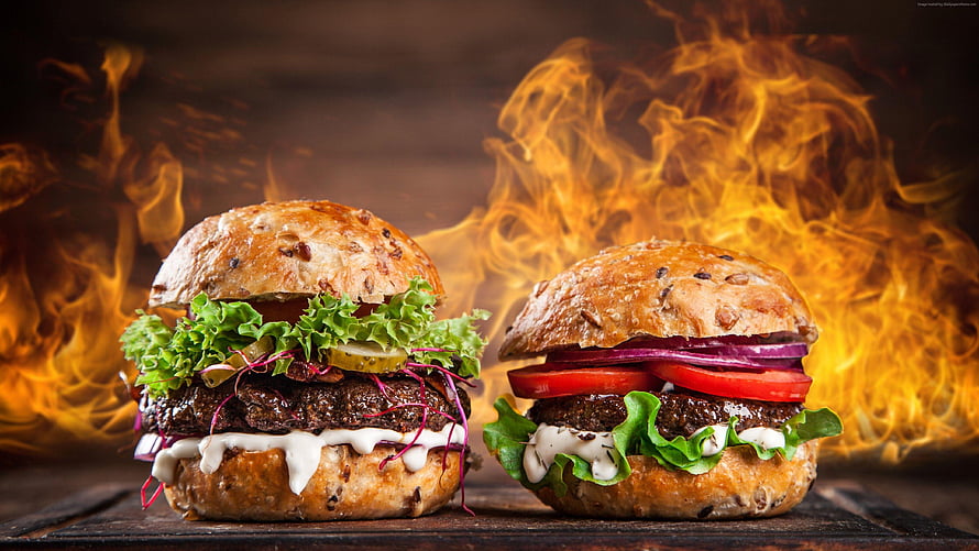 Two gourmet burgers with lettuce, tomato, and sauce set against dramatic flames HD wallpaper