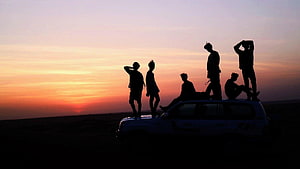 BTS Silhouettes on Car Roof at Sunset in Open Desert Landscape HD wallpaper