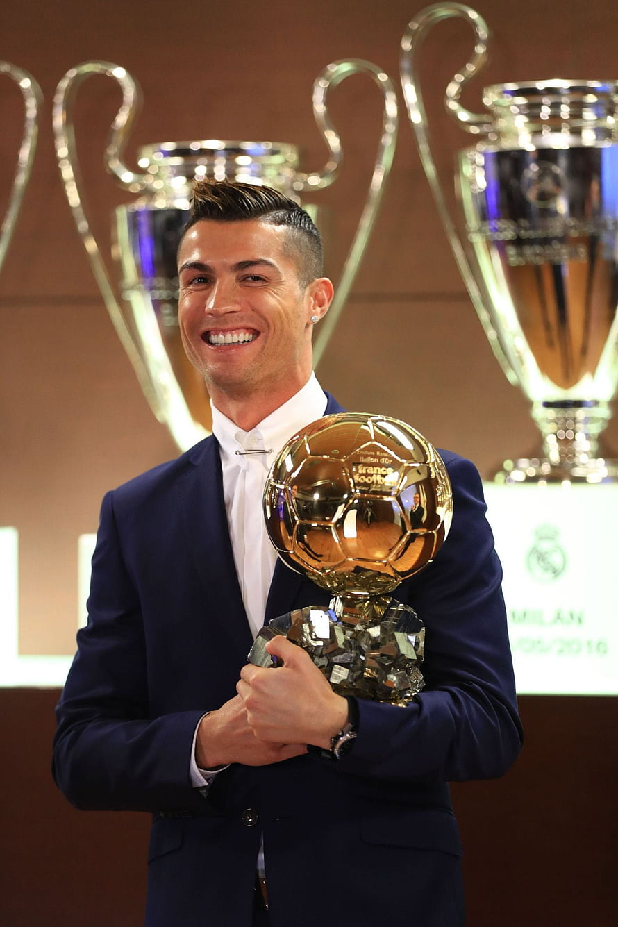Cristiano Ronaldo holding the Ballon d'Or trophy in a navy suit with Champions League cups in the background HD phone wallpaper