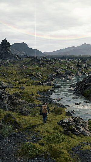 Hiker with backpack walking through mossy rocky landscape near river with rainbow in sky HD phone wallpaper