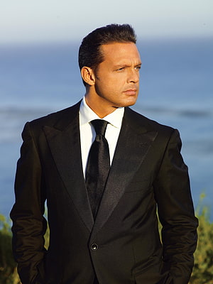 Luis Miguel in a black tuxedo with a serious expression against a blurred ocean backdrop HD phone wallpaper