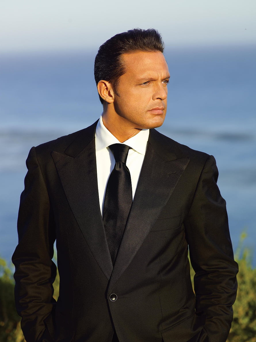 Luis Miguel in a black tuxedo with a serious expression against a blurred ocean backdrop HD phone wallpaper
