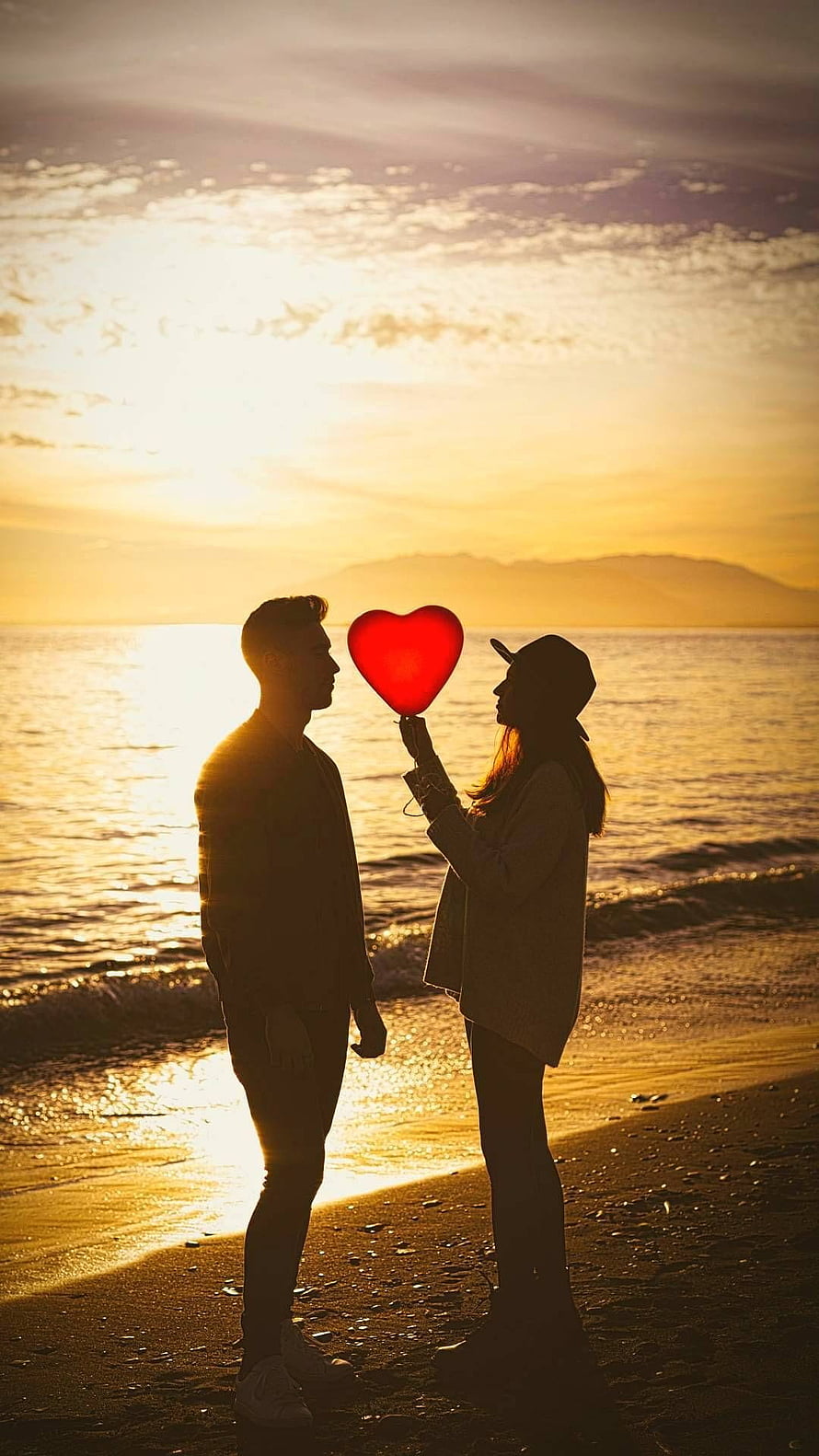 Silhouetted couple holding a red heart balloon on a beach at golden hour sunset HD phone wallpaper
