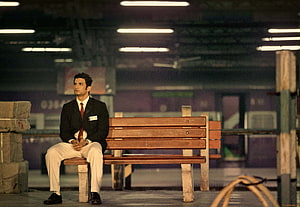 Sushant Singh Rajput as MS Dhoni in a formal suit sitting on a wooden bench in a dimly lit station HD wallpaper