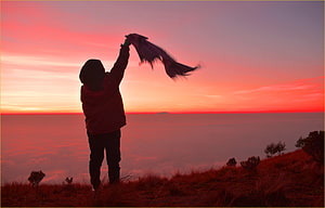 Silhouette of a person holding a scarf high on a mountain peak above a sea of clouds during a vibrant red sunset HD wallpaper