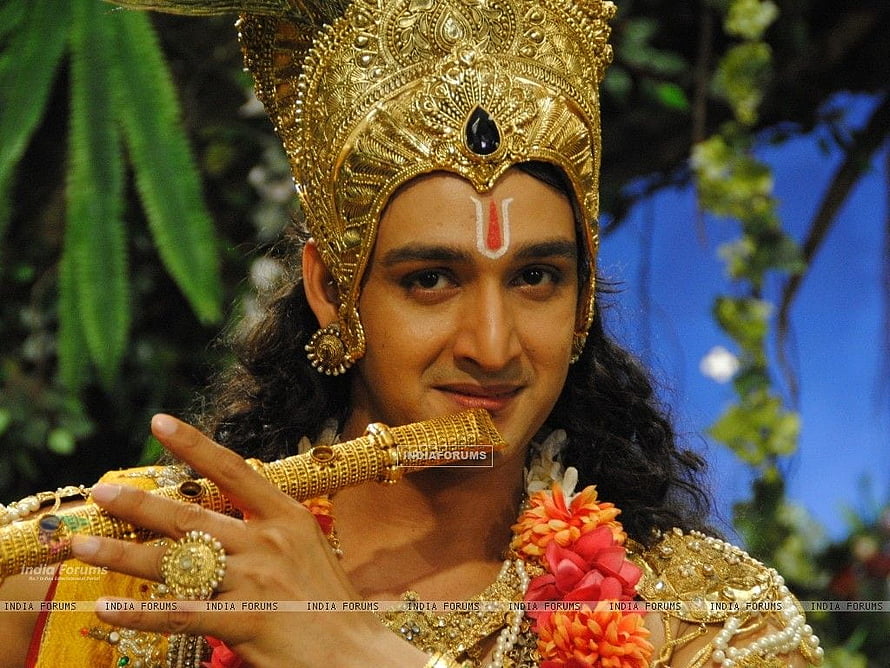 Krishna in ornate golden crown and jewelry holding a flute in a lush garden setting HD wallpaper