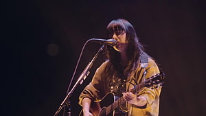 Aimyon performing live on stage with acoustic guitar and microphone under warm spotlight HD wallpaper
