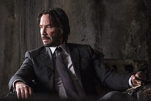 John Wick in a dark suit seated against a gritty concrete wall with a serious expression HD wallpaper