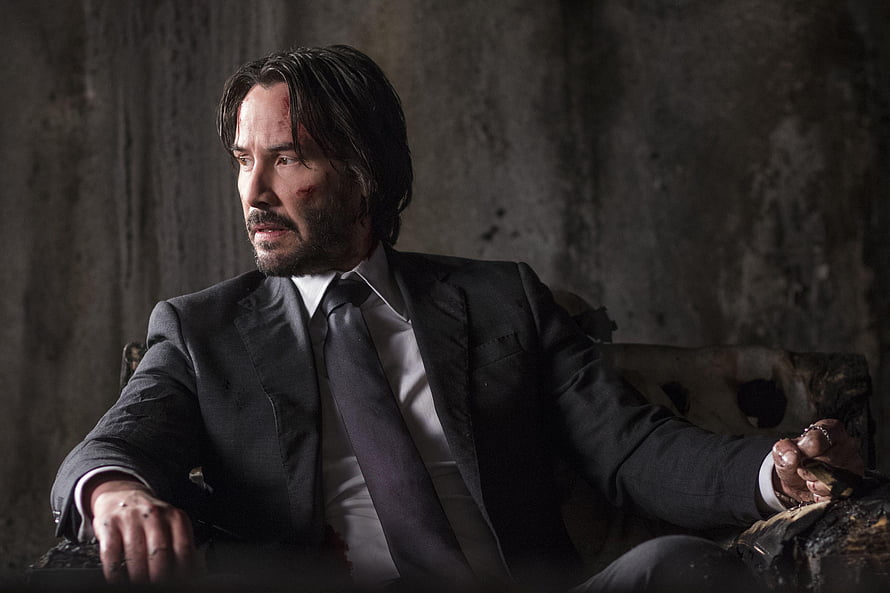 John Wick in a dark suit seated against a gritty concrete wall with a serious expression HD wallpaper