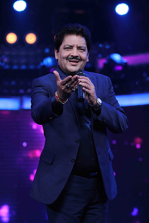 Udit Narayan performing on stage in a dark suit with microphone under blue and purple concert lighting HD phone wallpaper