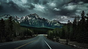 Moody Mountain Road Under Stormy Skies with Snow-Capped Peaks and Dense Forest HD wallpaper