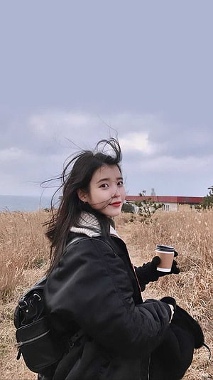IU in a black winter coat holding a coffee cup in a dry grassy field with a coastal backdrop HD phone wallpaper