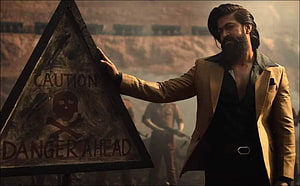 Yash in KGF Chapter 2 standing beside a caution danger sign in a dusty industrial setting HD wallpaper
