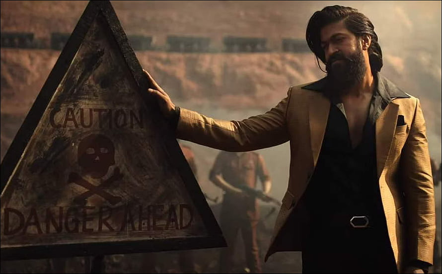 Yash in KGF Chapter 2 standing beside a caution danger sign in a dusty industrial setting HD wallpaper