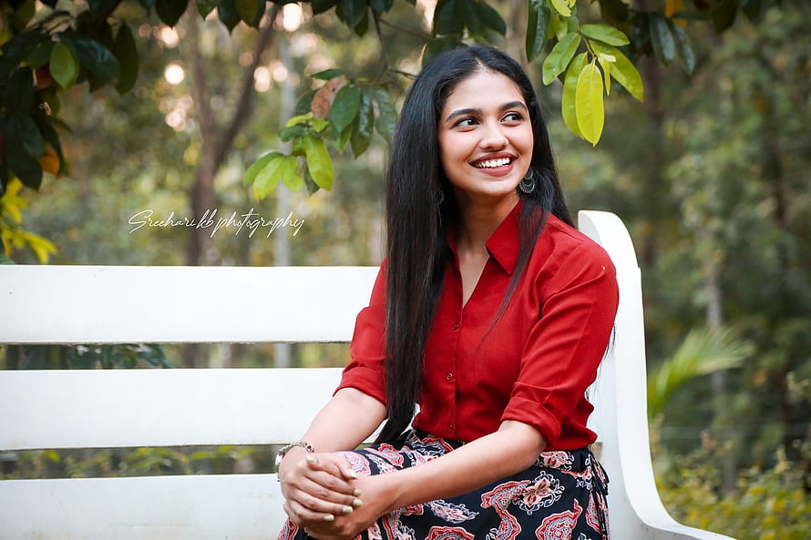 Mamitha Baiju smiling in a red shirt while seated on a white bench in a lush green park HD wallpaper