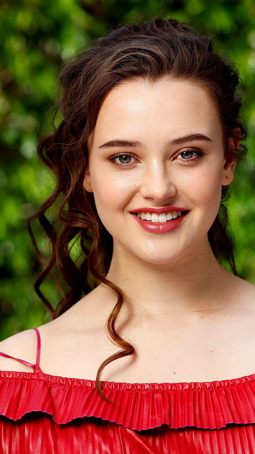 Katherine Langford smiling portrait in a red off-shoulder top with a lush green background HD phone wallpaper