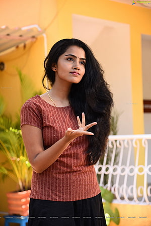 Portrait of a woman with long black hair wearing a rust-colored patterned top and black skirt, posing outdoors against a yellow wall, Anupama Parameswaran HD phone wallpaper