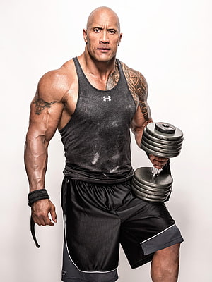 Dwayne Johnson in a black tank top and shorts holding a dumbbell with a focused expression HD phone wallpaper
