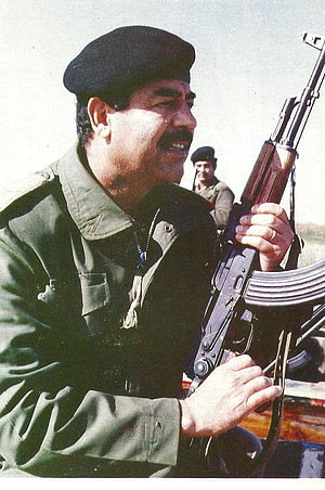 Saddam Hussein in military uniform holding an AK-47 rifle outdoors HD phone wallpaper
