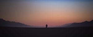 Silhouette of a lone figure on a motorcycle riding through a vast desert landscape at sunset with distant mountains HD wallpaper