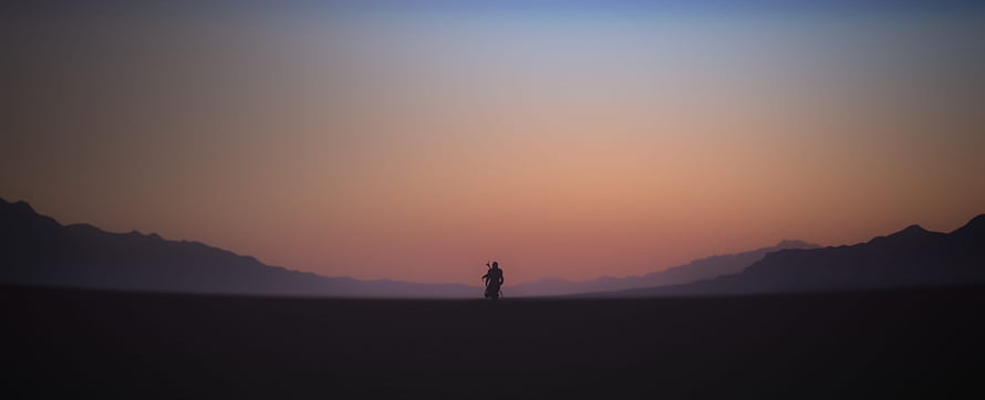 Silhouette of a lone figure on a motorcycle riding through a vast desert landscape at sunset with distant mountains HD wallpaper