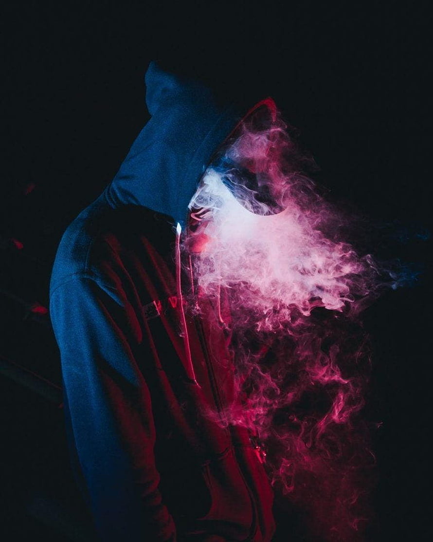 Mysterious hooded figure with swirling pink and blue smoke against a dark background HD phone wallpaper