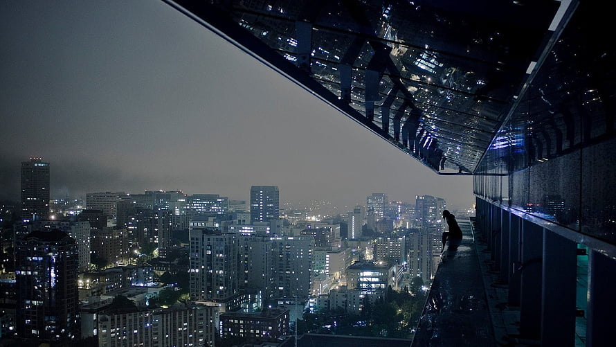Silhouette of a person sitting on a high-rise balcony overlooking a sprawling city skyline at night with glowing windows HD wallpaper